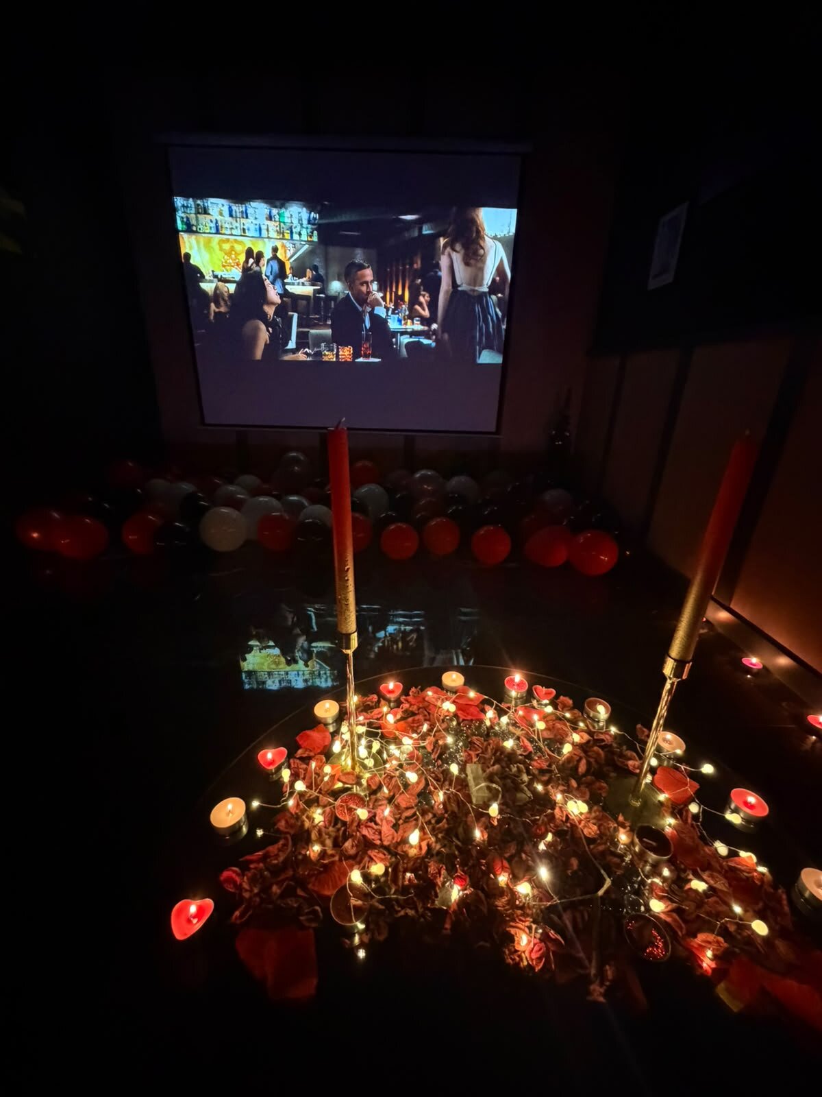 Perdede film oynarken önünde mumlar ve gül yapraklarıyla hazırlanmış kalp düzeni, arkada balonlar